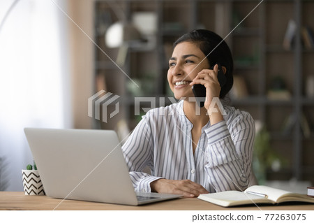 Smiling Indian woman talking on phone, sitting at work desk 77263075