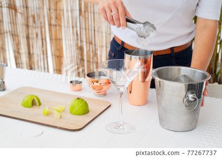 Bartender putting ice cube into shaker while making fresh cocktail with lime juice Bartender putting ice cube into shaker while making fresh cocktail with lime juice 77267737