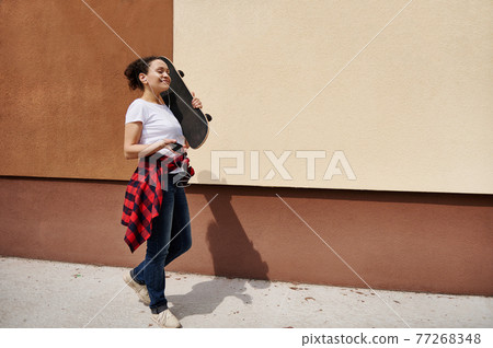 Happy woman holding wooden skateboard on shoulder walking down the street listening to music against colored wall background Happy woman holding wooden skateboard on shoulder walking down the street listening to music against colored wall background 77268348