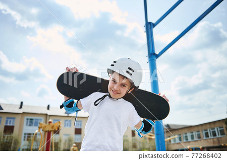 Adorable boy in a protective helmet and equipment posing with a skateboard on his shoulders behind his head 77268402