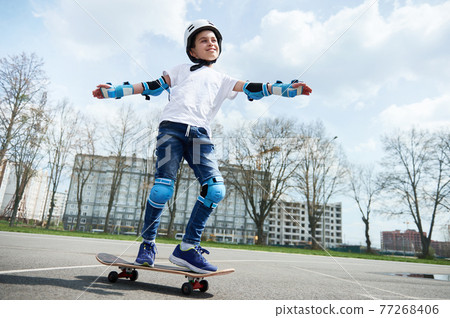 Happy and smiling boy in protective gear and helmet keeps balance while riding a skateboard 77268406