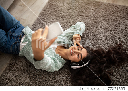 Above view of lovely Indian lady taking selfie while listening to music in headphones on floor at home Above view of lovely Indian lady taking selfie while listening to music in headphones on floor at home 77271865