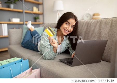 Beautiful Indian lady shopping online, lying on sofa with laptop pc and credit card, gift bags standing nearby 77271916