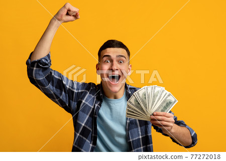 Overjoyed hipster guy holding lots of money, screaming WOW and making YES gesture on orange studio background 77272018