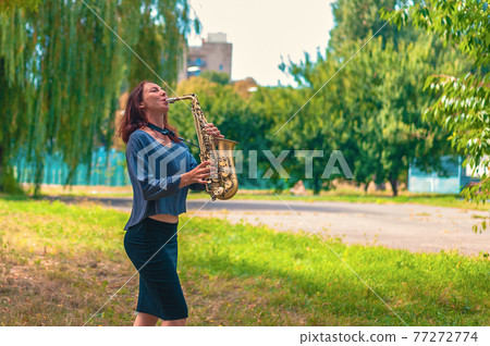 pretty young woman with red hair in a blue sweater and black skirt plays on a yellow saxophone in a city green park pretty young woman with red hair in a blue sweater and black skirt plays on a yellow saxophone in a city green park 77272774