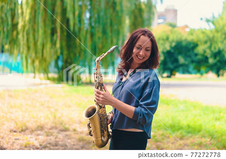 lovely girl with red hair holds a saxophone in her hands and laughs in nature 77272778