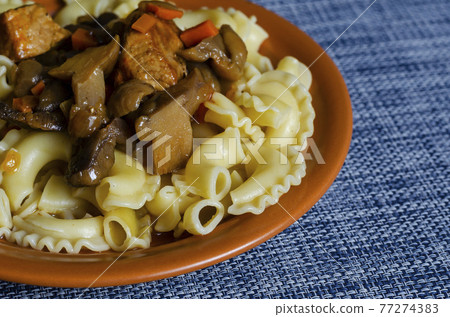 Serving of ready-to-eat pasta with sauce, stews, mushrooms, and vegetables in  clay plate on blue tablecloth. 77274383