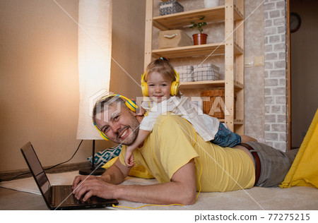 a little girl lies on her father's back and listens to music with yellow headphones while looking at her laptop. a little girl lies on her father's back and listens to music with yellow headphones while looking at her laptop. 77275215