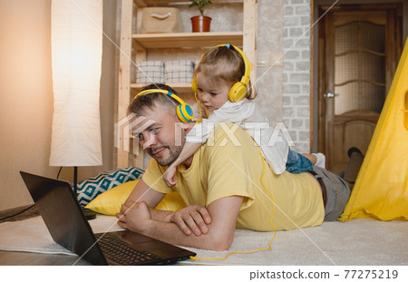 a little girl lies on her father's back and listens to music with yellow headphones while looking at her laptop. a little girl lies on her father's back and listens to music with yellow headphones while looking at her laptop. 77275219