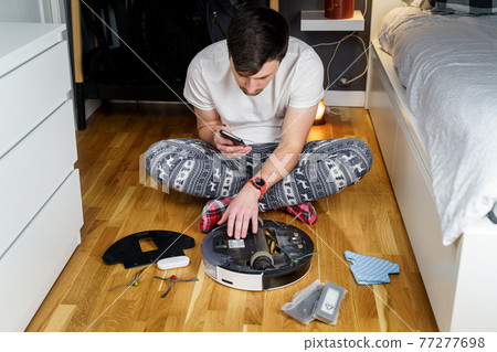 Caucasian young man in christmas pajamas doing vacuum cleaner robot maintenance. Male in winter patterned sleepwear and red socks slippers starts automatic vacuum cleaner. Robot vacuum cleaner repair Caucasian young man in christmas pajamas doing vacuum cleaner robot maintenance. Male in winter patterned sleepwear and red socks slippers starts automatic vacuum cleaner. Robot vacuum cleaner repair 77277698