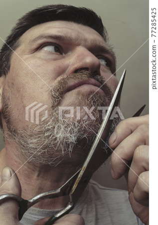 An adult man cuts his own beard with old huge scissors. An adult man cuts his own beard with old huge scissors. 77285425