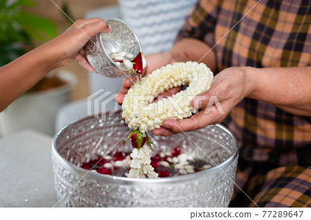 Point of view of child pouring water on hands of elder senior or respected grandparents who holding jasmine garland on hand for celebrate Songkran in new year water festival. 77289647