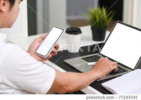 Close up view young man entrepreneur holding smart phone and working with laptop computer at modern workplace. 77293595