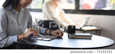 Cropped shot of happy female employee working on digital tablet while sitting with her colleague in office. 77293783