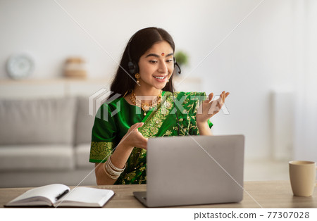 Positive Indian woman in traditional sari dress wearing headphones, having online meeting on laptop at home Positive Indian woman in traditional sari dress wearing headphones, having online meeting on laptop at home 77307028