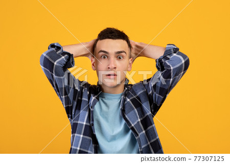 Emotional young man grabbing his head in shock, looking at camera on orange studio background. Human emotions Emotional young man grabbing his head in shock, looking at camera on orange studio background. Human emotions 77307125