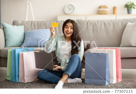 Indian woman feeling happy over sale in online shop, using laptop, showing credit card, sitting on floor with gift bags Indian woman feeling happy over sale in online shop, using laptop, showing credit card, sitting on floor with gift bags 77307215