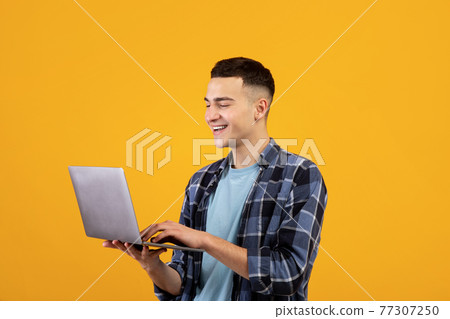 Happy young man looking at laptop screen with smile, watching interesting web content on orange studio background Happy young man looking at laptop screen with smile, watching interesting web content on orange studio background 77307250