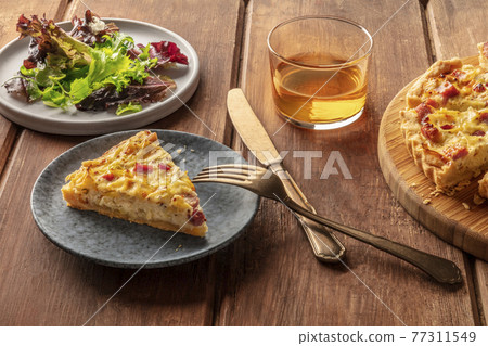A French dinner. A slice of a quiche lorraine with mesclun leaves and white wine on a rustic wooden background 77311549