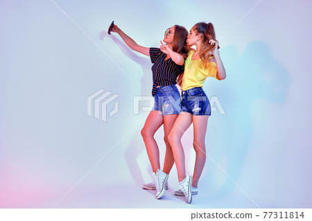 Two teenage girls in summer clothes standing in studio and making selfie on white background. Mixed light Two teenage girls in summer clothes standing in studio and making selfie on white background. Mixed light 77311814