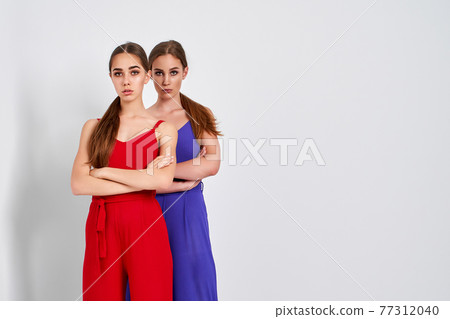 Two girls standing in studio in elegant colorful overalls on white background Two girls standing in studio in elegant colorful overalls on white background 77312040