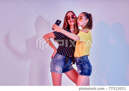 Two teenage girls in summer clothes standing in studio and making selfie on white background. Mixed light Two teenage girls in summer clothes standing in studio and making selfie on white background. Mixed light 77312051