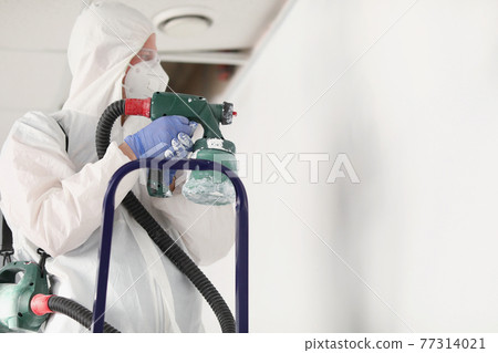 Male painter in protective suit is painting wall on stepladder with spray gun 77314021