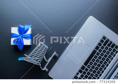 Web shopping. Laptop computer, shopping trolley and white gift with blue ribbon on dark background. Ecommerce and customer experience concept. Internet purchase, online shop. 77314209