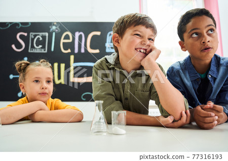 Group of multiracial kids sitting at the desk and listening their teacher Group of multiracial kids sitting at the desk and listening their teacher 77316193