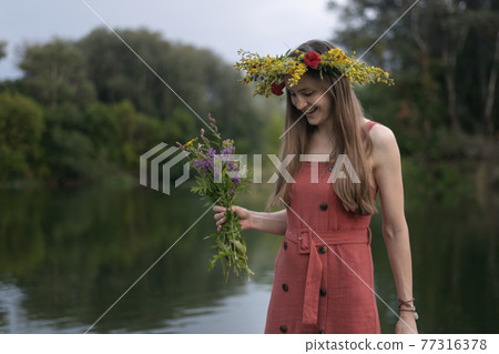 Young woman with wreath on her head with bouquet of wildflowers. River and forest on background. Summer solstice 77316378