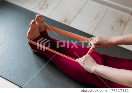 Resistance band exercise at home. Woman doing pilates workout using elastic strap pulling with arms for shoulder training on yoga mat indoors. Sport and healthy active lifestyle concept. Resistance band exercise at home. Woman doing pilates workout using elastic strap pulling with arms for shoulder training on yoga mat indoors. Sport and healthy active lifestyle concept. 77317511
