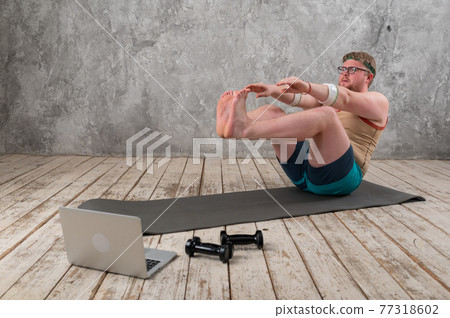 funny fat man in sports clothes doing exercises. He plays sports remotely while looking at his laptop 77318602