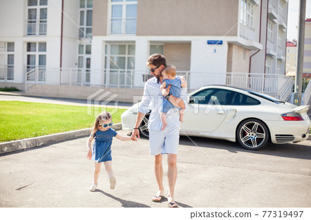 young dad walks with his two daughters on the background of a sports car young dad walks with his two daughters on the background of a sports car 77319497