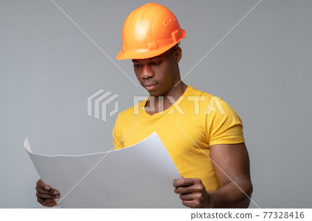 Serious man in helmet considering blueprint drawing Serious man in helmet considering blueprint drawing 77328416