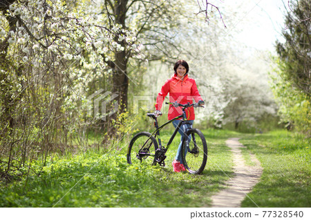 Beautiful woman on a bike in a blooming spring garden. Beautiful mature woman posing for the camera in a blooming spring garden. Beautiful woman on a bike in a blooming spring garden. Beautiful mature woman posing for the camera in a blooming spring garden. 77328540