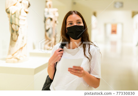 Young girl wearing a protective mask in the museum looks at the exhibits 77330830