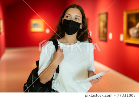 Young girl in a protective mask visits an exhibition of paintings in a museum 77331005