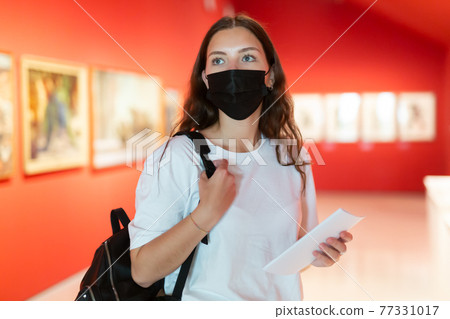 Young girl in a protective mask visits an exhibition of paintings in a museum 77331017