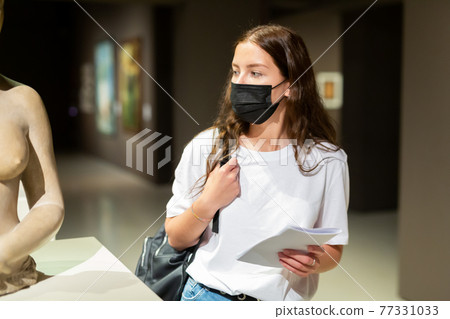 Portrait of a young girl visitor in a protective mask, looking at a sculpture in the museum hall 77331033