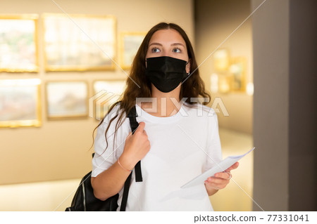 Young girl in a protective mask visits an exhibition of paintings in a museum 77331041