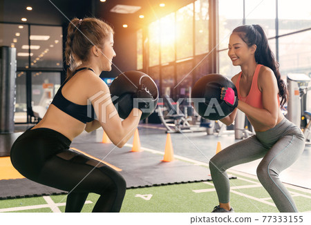 Happy young female athletic people performing squat exercises with friend and holding a medicine ball at fitness gym. Group of two confident women with healthy lifestyle working out together. Happy young female athletic people performing squat exercises with friend and holding a medicine ball at fitness gym. Group of two confident women with healthy lifestyle working out together. 77333155