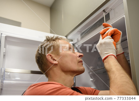 The worker is fixing the built-in refrigerator in the kitchen furniture. 77333844