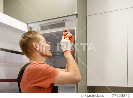 The worker is fixing the built-in refrigerator in the kitchen furniture. 77333845