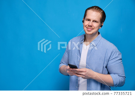 A man in a blue shirt dressed wireless headphones holds a smartphone and looks at the camera. The concept of ease of communication and modern technology. A man in a blue shirt dressed wireless headphones holds a smartphone and looks at the camera. The concept of ease of communication and modern technology. 77335721