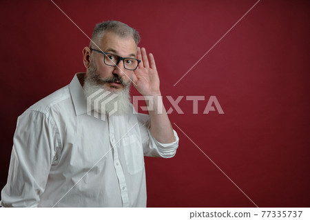 Mature bearded man dressed white shirt holds hand near ear, want to know information, red background 77335737