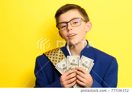 A boy in glasses and an oversized suit with a satisfied face is holding US dollars, a child and big money. A boy in glasses and an oversized suit with a satisfied face is holding US dollars, a child and big money. 77336152