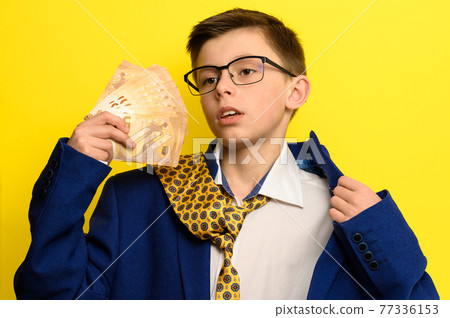 A child with a pathetic look holds in his hands euro bills, a boy in an adult suit on a yellow background, a blue jacket and a yellow tie. A child with a pathetic look holds in his hands euro bills, a boy in an adult suit on a yellow background, a blue jacket and a yellow tie. 77336153