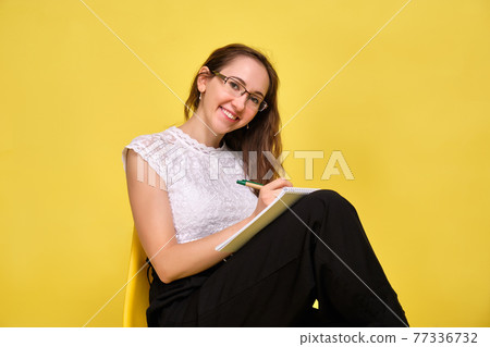 Girl smiling teacher in glasses on a yellow background sits and writes a pen in a notebook. 77336732