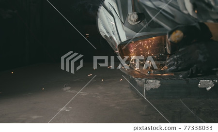 Forge workshop. Smithy. Worker in a welding hood helmet welds a part by electric welding. Sparks are reflected in the protective screen. 77338033