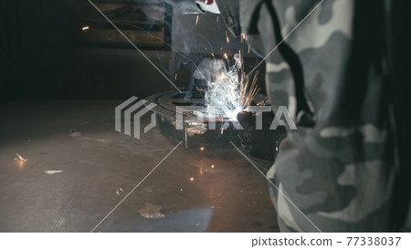 Forge workshop. Smithy. Worker in a welding hood helmet welds a part by electric welding. Sparks are reflected in the protective screen. Forge workshop. Smithy. Worker in a welding hood helmet welds a part by electric welding. Sparks are reflected in the protective screen. 77338037